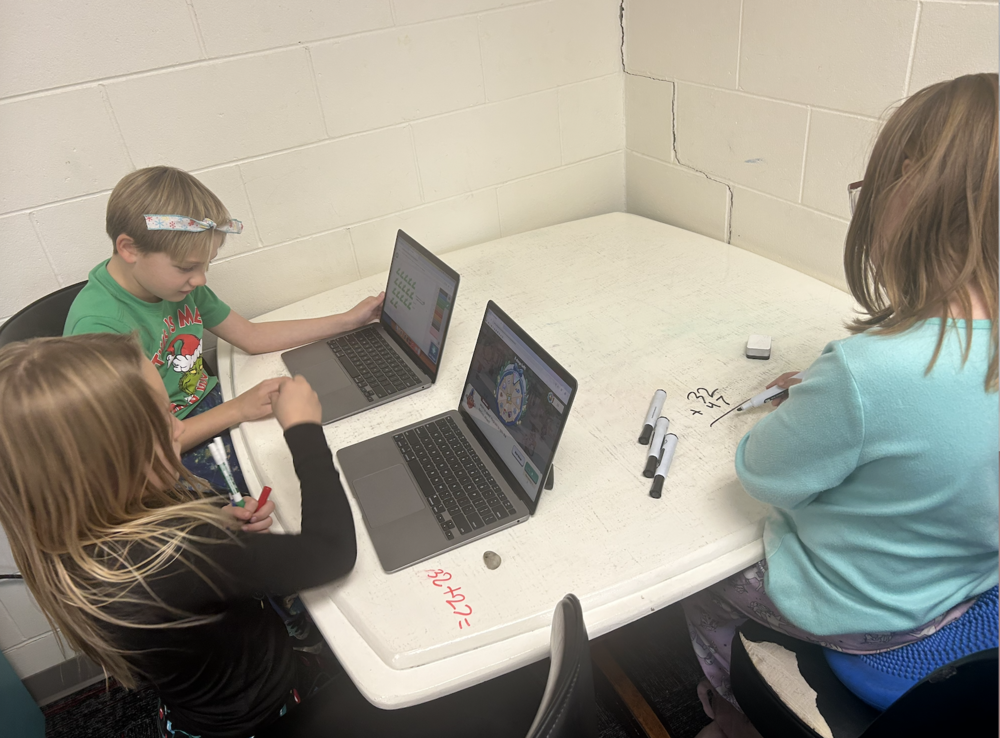 Three elementary students sit around a white table working collaboratively with two open laptops. One student uses a dry-erase marker to write on the tabletop while the others look at coding or interactive activities on their screens. Dry-erase markers and laptops are spread across the table, showing hands-on learning and teamwork in a classroom setting.