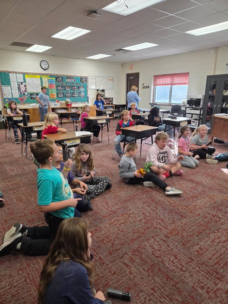 Elementary students sit on the carpet and at their desks in a classroom, watching something toward the front of the room. Some hold small objects or water bottles, and a teacher stands near the back. The classroom has bulletin boards, desks, bright lighting, and a large window letting in natural light.