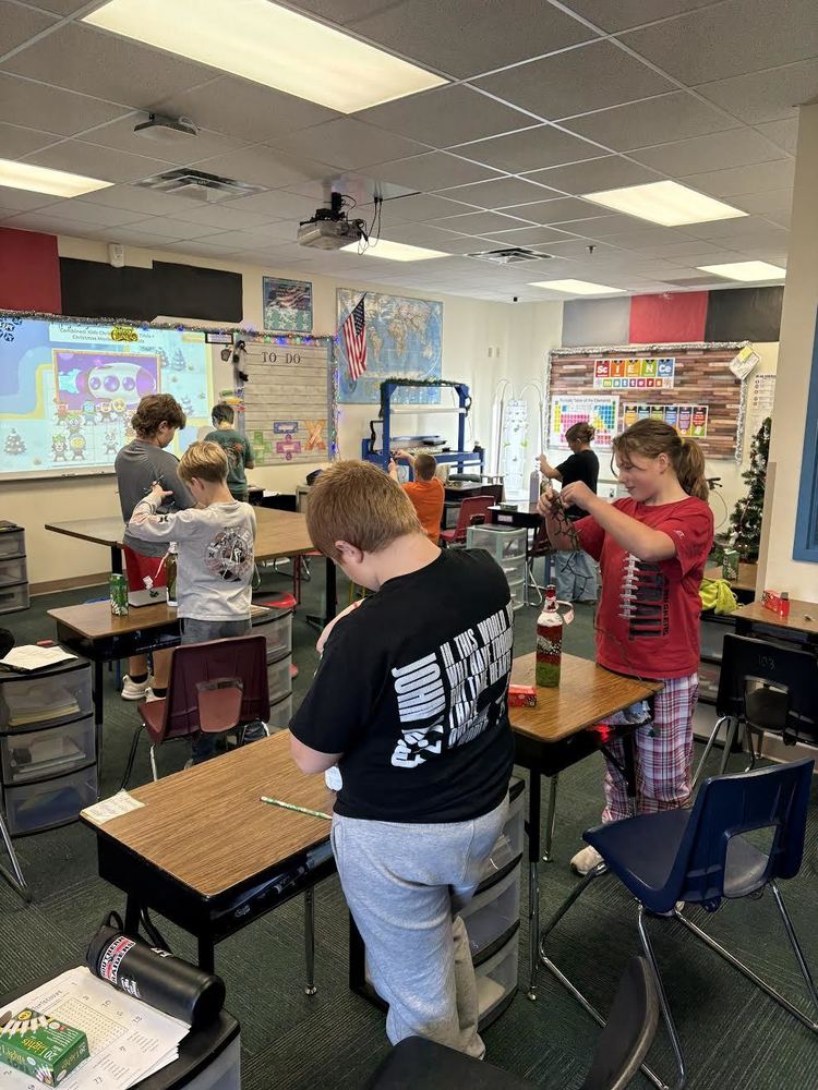 Elementary students stand at their desks working on a hands-on classroom activity, carefully assembling holiday-themed projects using small materials during a festive learning activity.