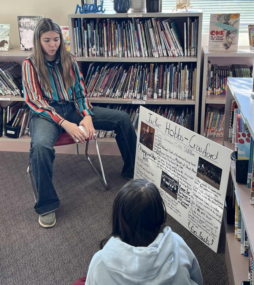 A student sits on a chair in the school library presenting a biography project about Jackie Hobbs-Crawford. Another student sits on the floor listening. A large poster board with photos, facts, and handwritten notes is displayed beside them, with bookshelves full of children’s books in the background.