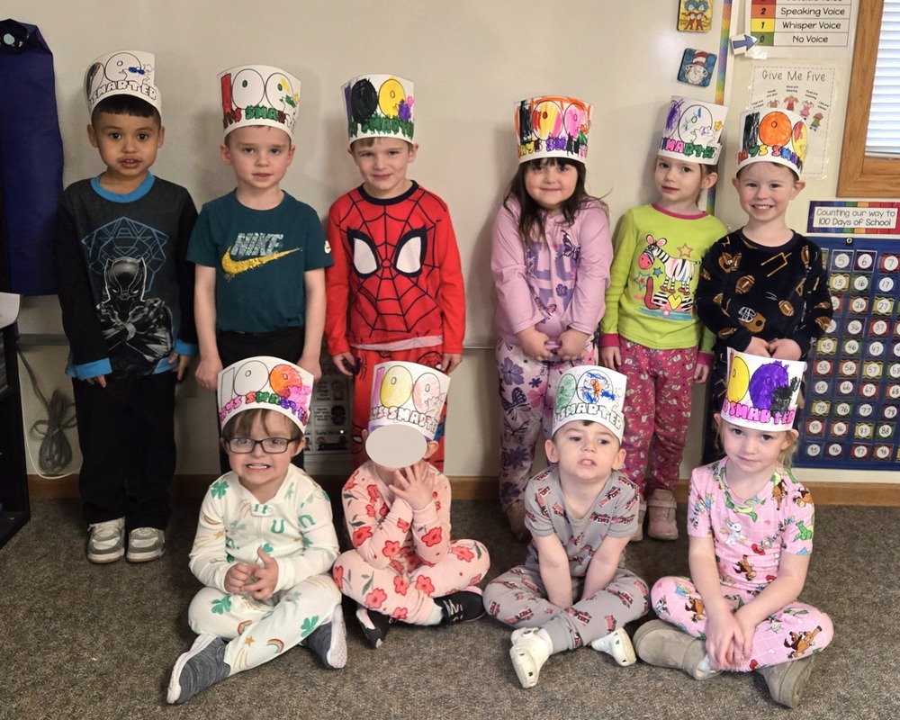 Ten preschool children pose together in a classroom, wearing colorful handmade paper crowns decorated with “100” to celebrate 100 days of school. Five children stand in the back row and five sit on the floor in front. They are dressed in pajamas and casual clothes, smiling or looking at the camera. A classroom wall with posters and a counting chart is visible behind them.