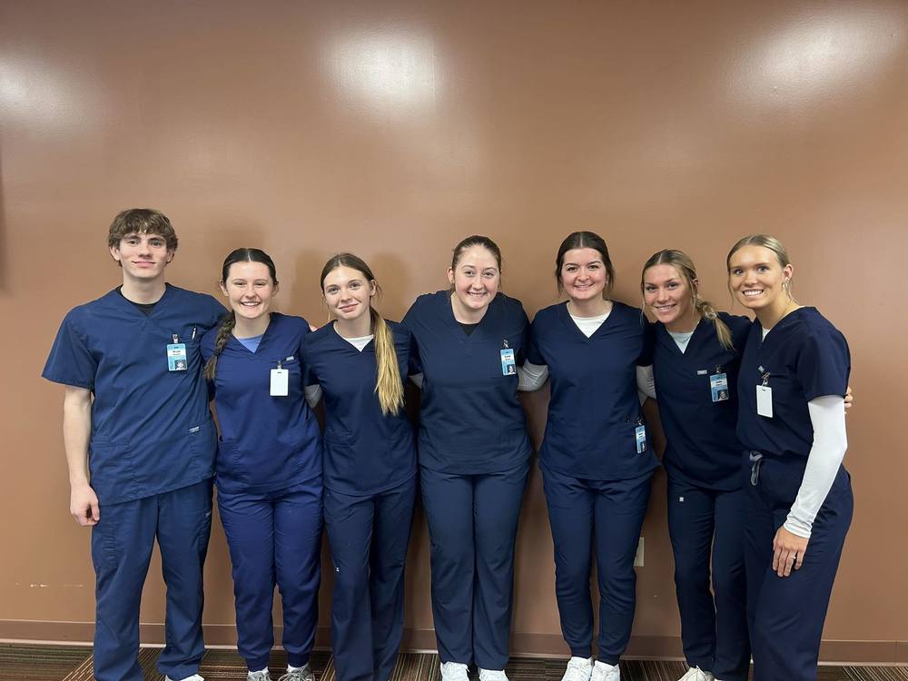 7 CNA students dressed in blue scrubs are standing in front of an orange background