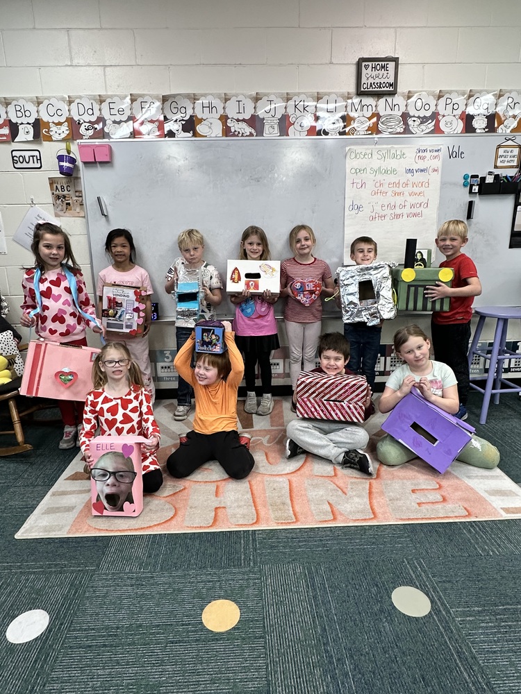 A group of about a dozen young children in an elementary classroom pose together on a rug that says “SHINE,” each holding a decorated cardboard box project. They stand and sit in two rows in front of a whiteboard and an alphabet border, smiling and showing their colorful creations made with paper, foil, and drawings.