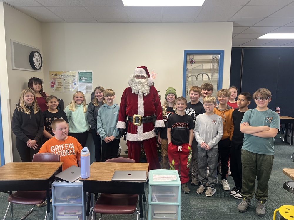 A group of elementary students stand together in a classroom smiling for a photo with a person dressed as Santa Claus, celebrating a festive holiday visit during the school day.