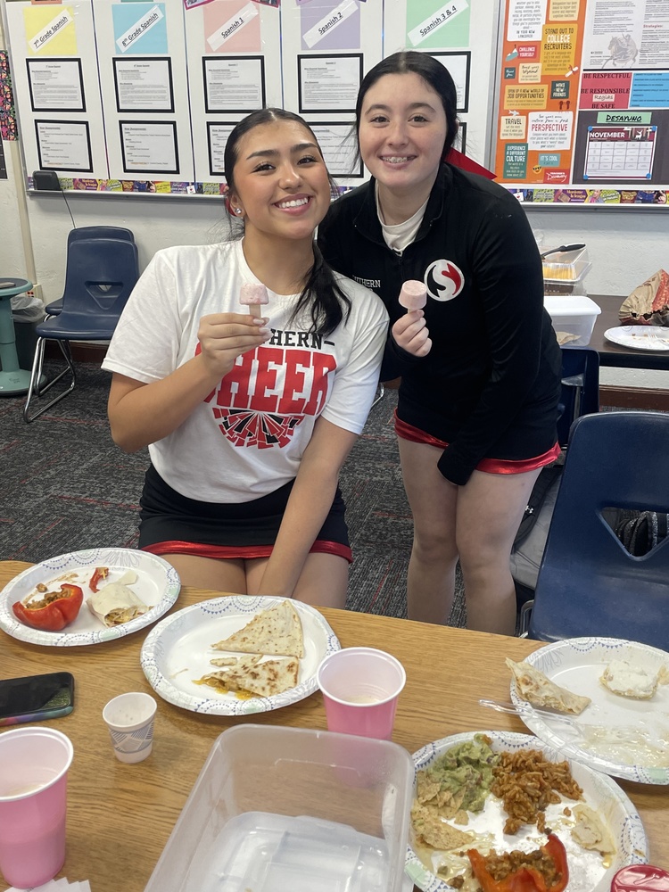Two cheerleaders smile while sitting at a classroom table, each holding a small frozen treat on a stick. Plates of food, cups, and snacks are spread out on the table in front of them, suggesting a class celebration or team meal.
