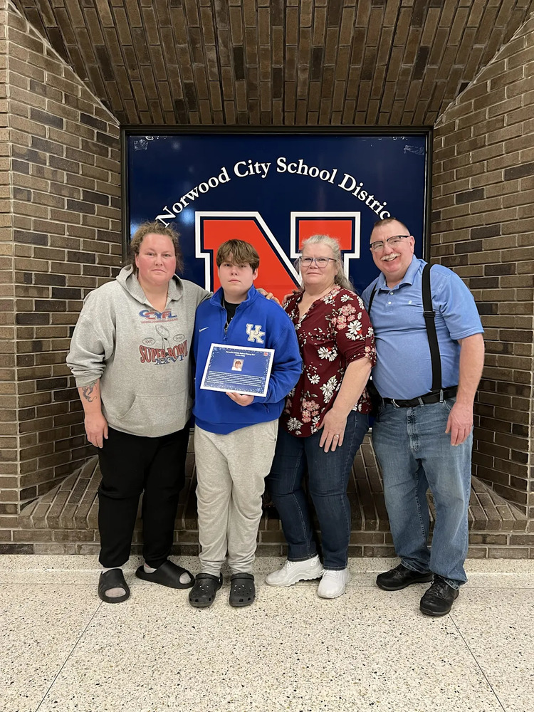 Four people stand together indoors in front of a sign reading “Norwood City School District” with a large red “N” behind them. A teenage boy in the center holds a certificate and wears a blue jacket and light-colored pants. Two women stand on either side of him, and a man stands on the far right. They are posing closely together against a brick wall background, a school recognition or award moment.