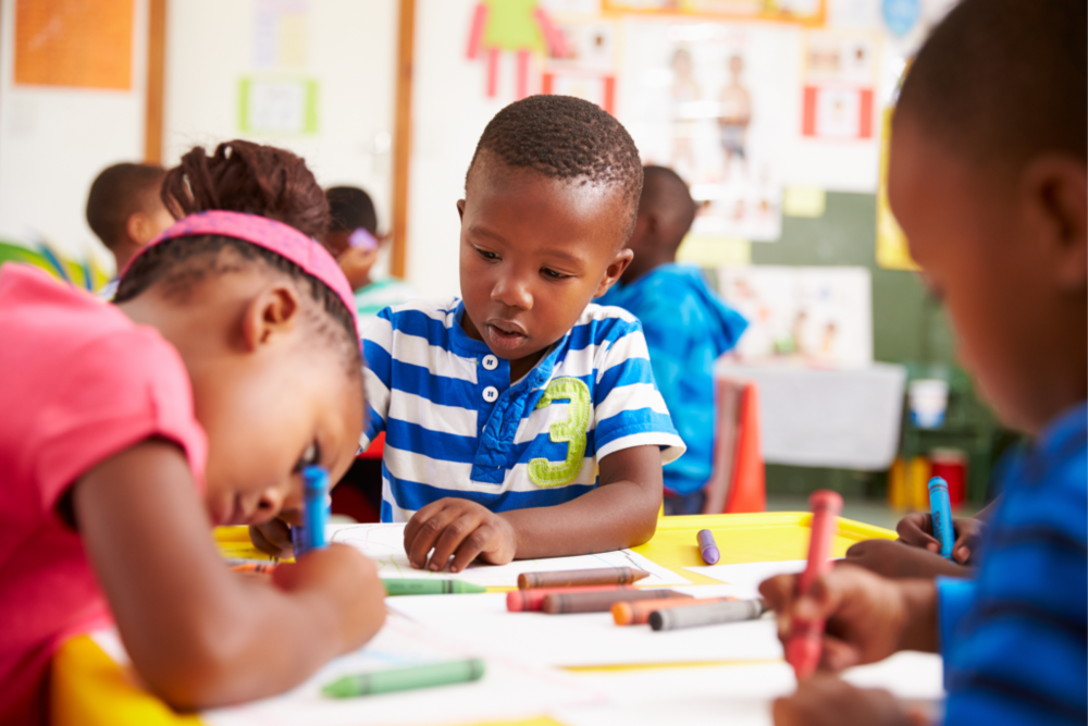 Young students sit around a classroom table drawing with crayons, concentrating on their artwork during a classroom activity.