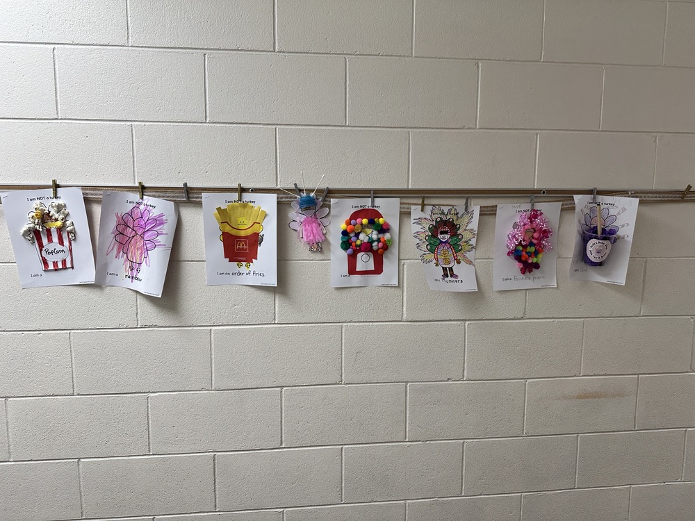 A row of children’s “I Am Not a Turkey” art projects hangs on a wall, each clipped to a horizontal display bar. The papers show creatively disguised turkeys, including designs like popcorn, a flower, French fries, a gumball machine, a ballerina, and other colorful, decorated characters. The wall behind them is light-colored brick.