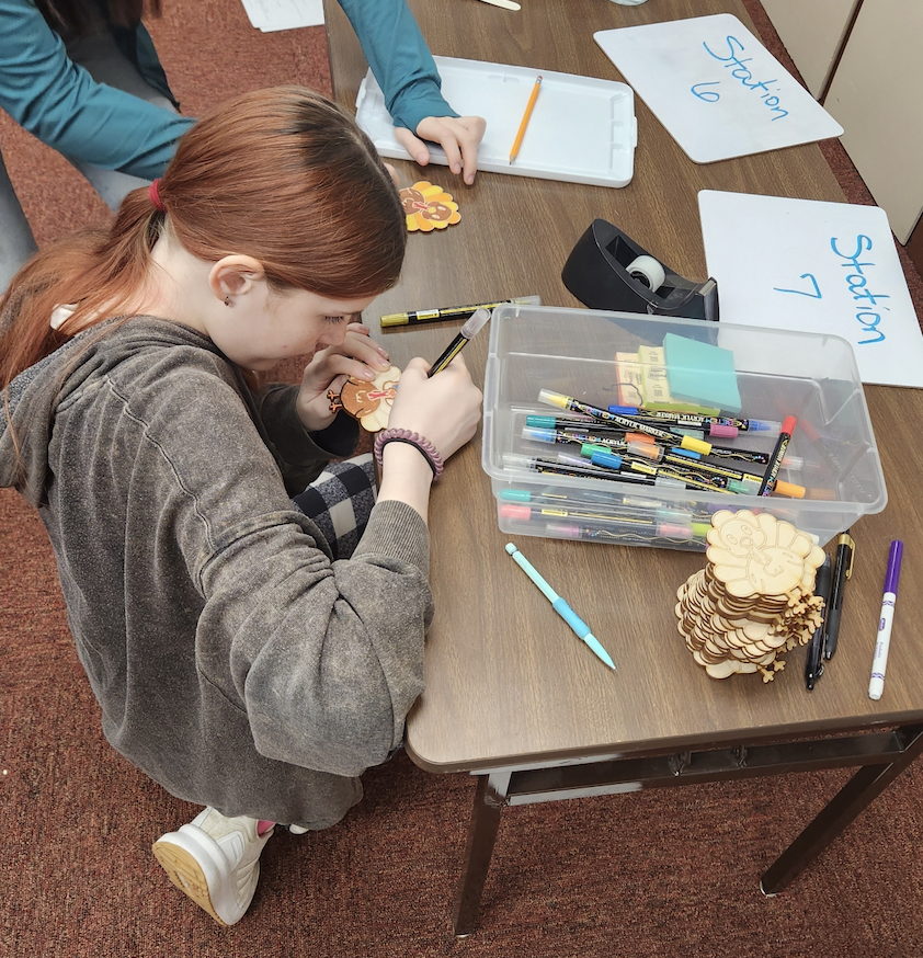 A student kneels beside a classroom table, carefully coloring a small wooden turkey craft using markers. A plastic bin filled with various markers sits on the table, along with a tall stack of unfinished wooden turkey pieces and whiteboards labeled “Station 6” and “Station 7.” Another student’s hands are visible nearby, also working on a craft.