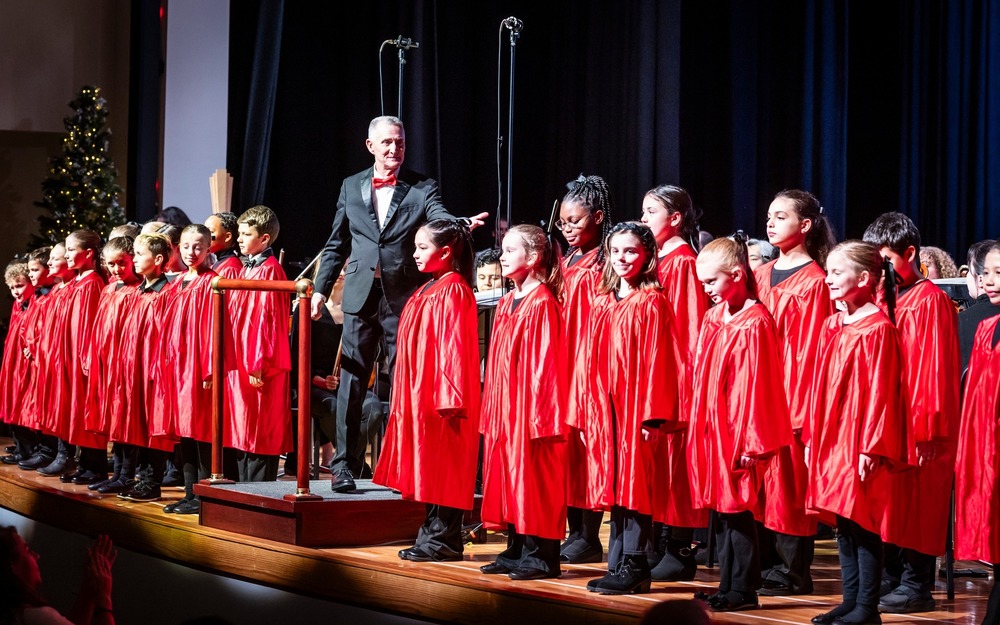 A group of children in red gowns with an adult in a black suit on a stage performing.