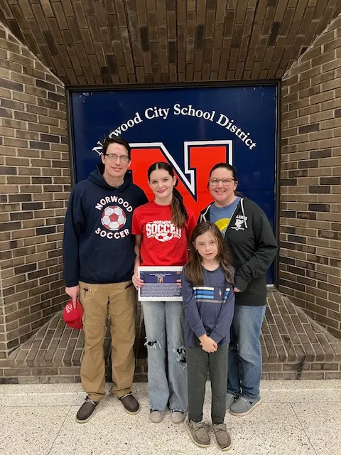 Four people pose together indoors in front of a “Norwood City School District” sign with a large red “N.” A teenage girl in the center holds a certificate and wears a red Norwood Soccer shirt and jeans. To her left stands a teenage man in glasses wearing a dark Norwood Soccer hoodie and khaki pants. To her right is an adult woman in glasses wearing a dark jacket. A younger girl stands in front, wearing a dark long-sleeve shirt and pants. The group stands close together against a brick wall background, a school recognition or award moment.