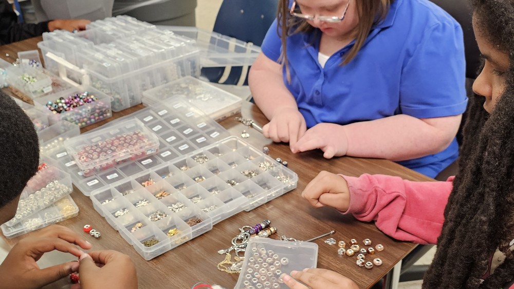 Students seated at a table selecting beads from small containers and creating jewelry during a classroom activity.