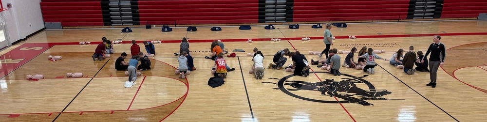 Students practice CPR in small groups on training mannequins spread across the gym floor during a hands-on safety and emergency response lesson