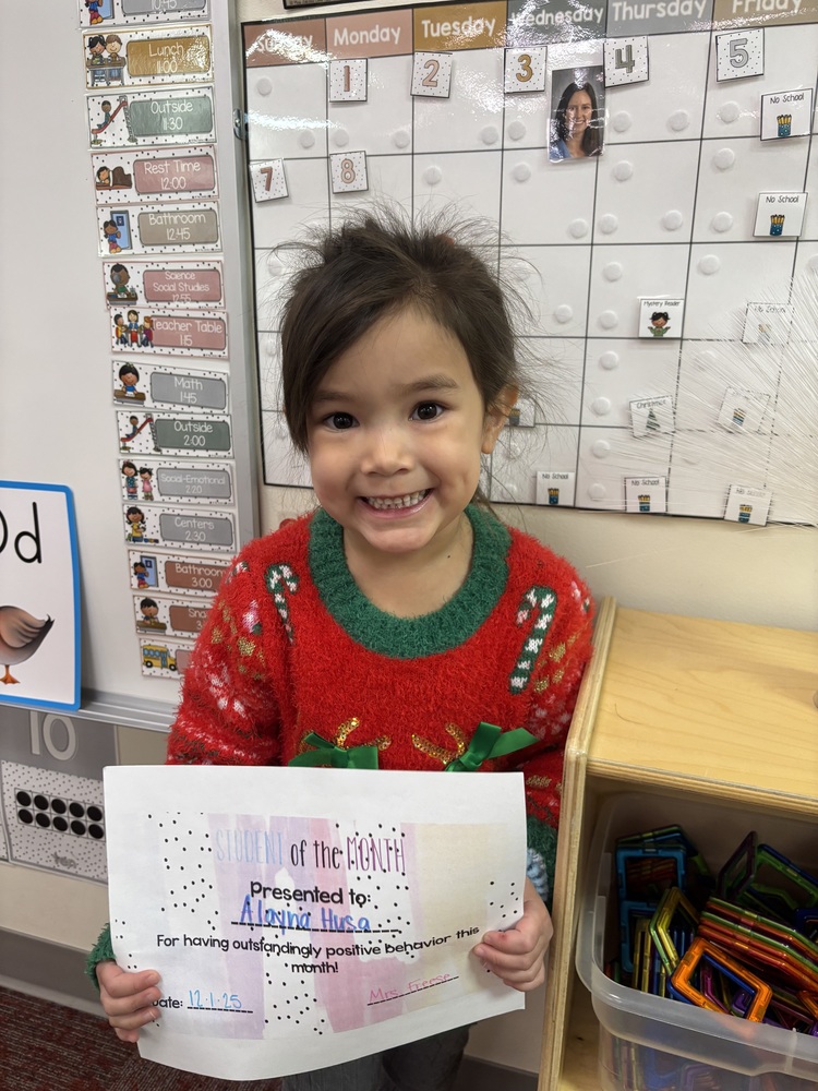 A smiling elementary student wearing a red holiday sweater stands in a classroom holding a “Student of the Month” certificate. The student is posing proudly in front of a classroom calendar and learning materials, celebrating recognition for positive behavior.