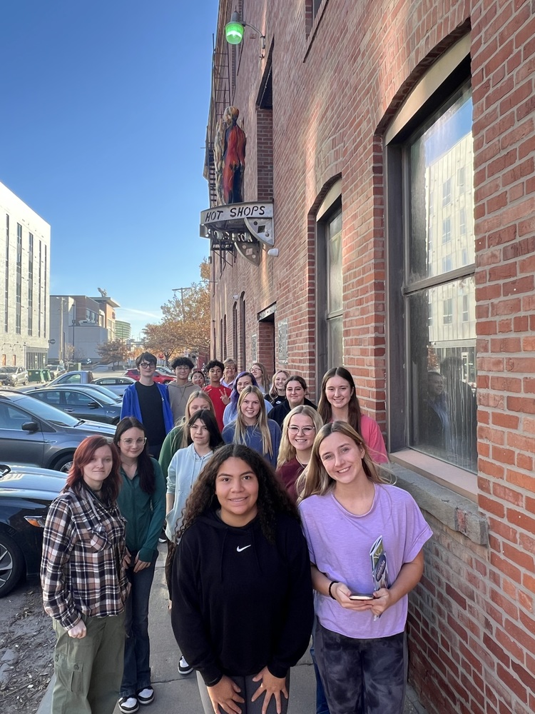A group of high school students stand in a line along a brick building on a sunny day outside Hot Shops Art Center. The students smile toward the camera, with parked cars and city buildings visible in the background.