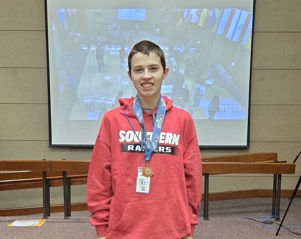 A student wearing a red Southern Raiders hoodie and black pants stands indoors, smiling and holding a medal on a blue lanyard, with a presentation screen and flags visible in the background.