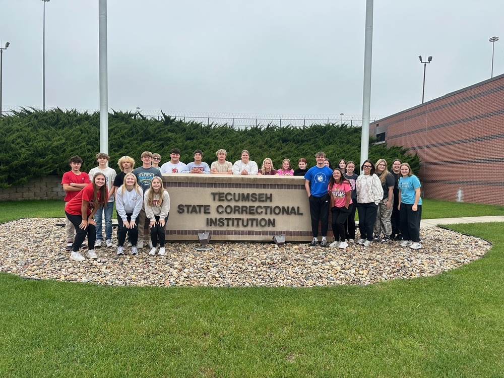 A group of high school students and staff pose outdoors in front of a stone sign reading “Tecumseh State Correctional Institution.” The group stands on grass and landscaping rocks, with the facility buildings and tall light poles visible in the background.