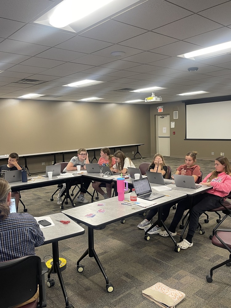 Students sit around tables in a classroom working on laptops, reviewing papers, and collaborating during a group learning session.