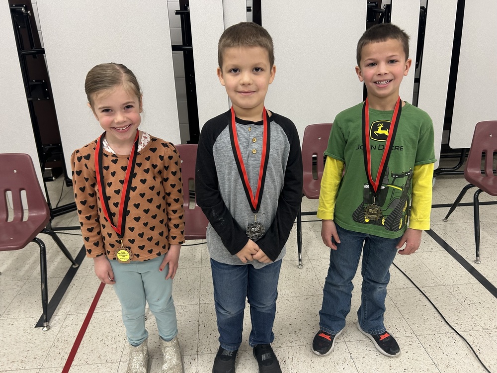 Three smiling children stand in a row, each wearing a medal on a red ribbon. They are in a room with maroon chairs, creating a joyful, celebratory mood.