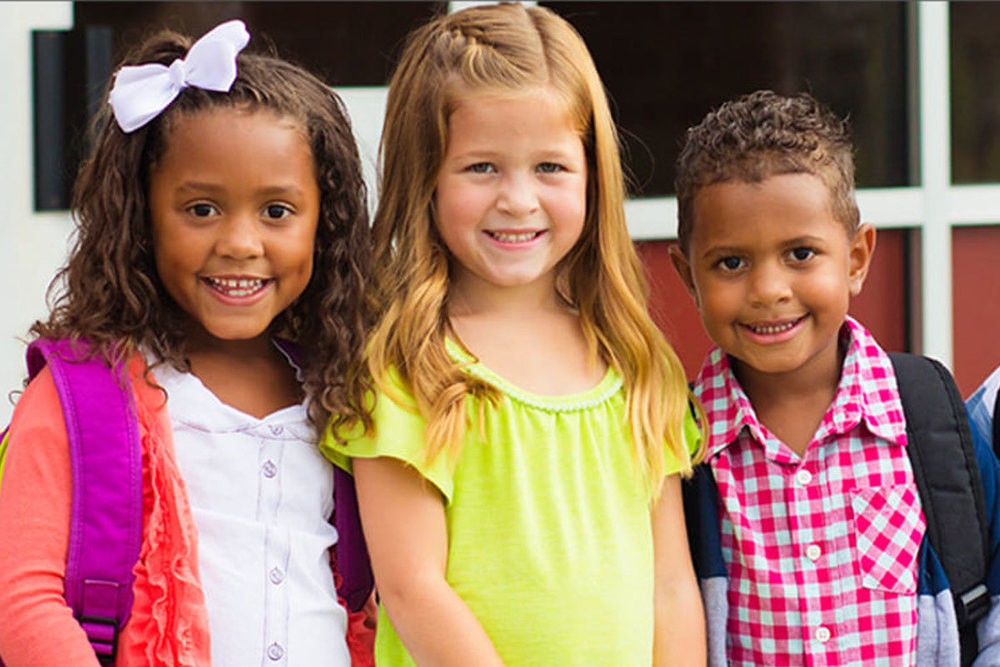 Three elementary school students wearing backpacks stand together outside a school building and smile for the camera.
