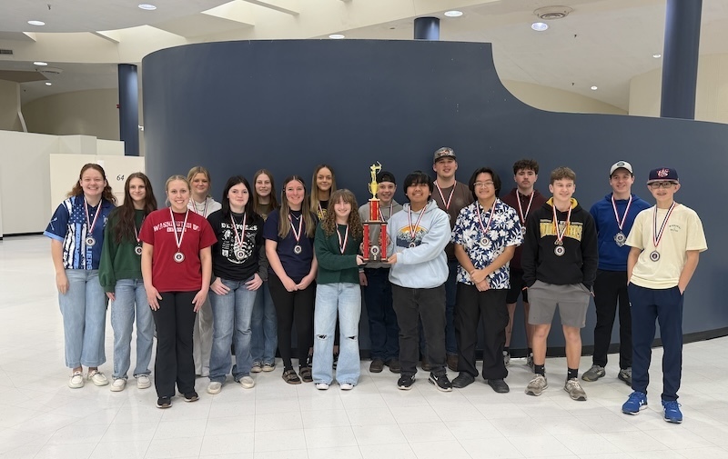 teenage aged students posing in front of a curved blue wall with a trophy and medals around their necks 