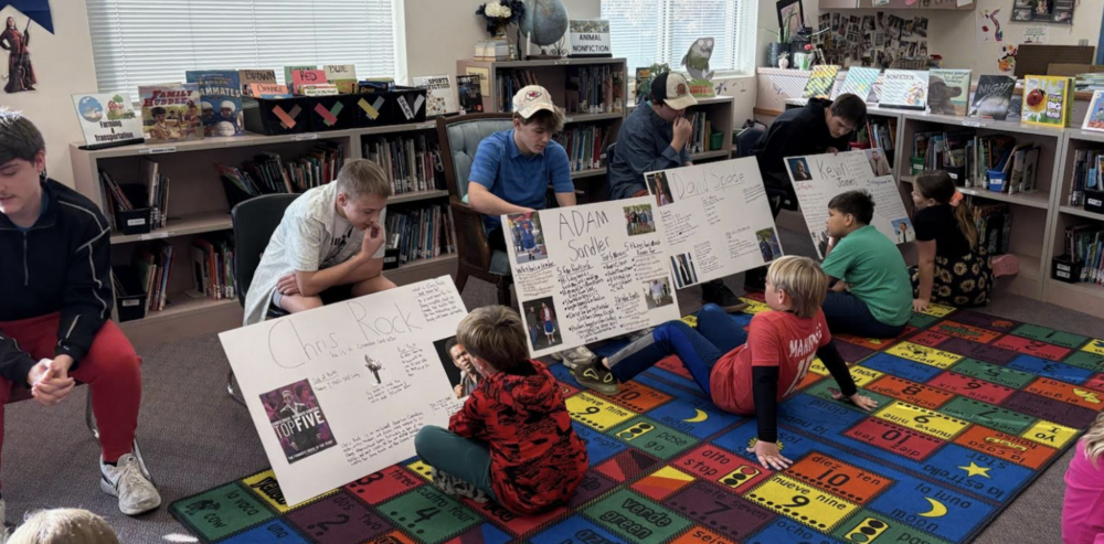 Older students sit in a library presenting large biography posters to younger children seated on a colorful alphabet-and-numbers rug. Each poster features a different comedian or actor with photos and handwritten facts. Bookshelves filled with children’s books line the background, and sunlight comes through the windows above them.