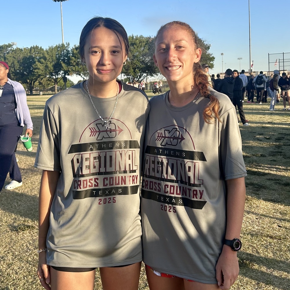 Hannah Hunt and Donna Carpenter smile for a photo after the UIL Regional Cross Country Meet in Arlington. Both are wearing matching gray Athens Regional Cross Country 2025 shirts, standing on a grassy field with other runners and spectators in the background under a clear sky.