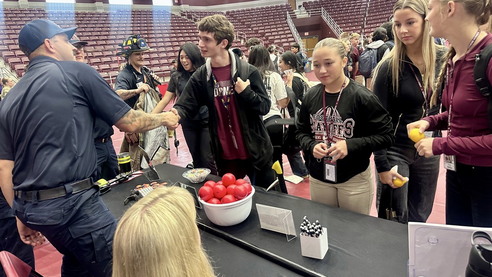 Athens High School students visit with members of the Athens Fire Department during the Henderson County Career Expo. One student shakes hands with a firefighter across a table displaying fire safety tools, red stress balls, and pens. Other students stand nearby holding giveaway items and listening. The event is taking place inside the AHS gymnasium.