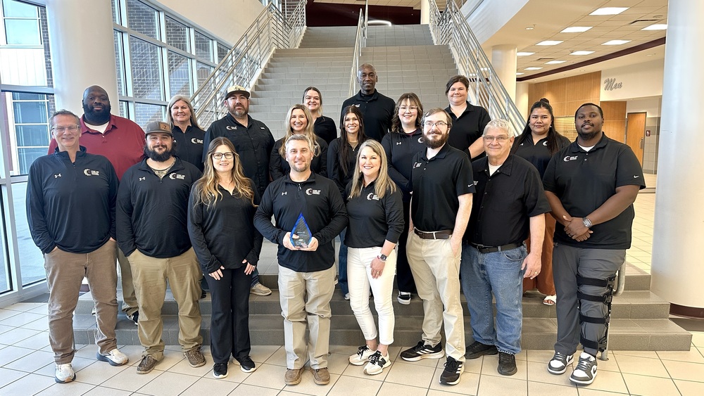 Group photo of Athens ISD Career and Technical Education staff standing on a wide indoor staircase inside a school building, wearing black Athens ISD CTE shirts. One staff member in the front center holds a glass award recognizing the district as a CTE District of Distinction.