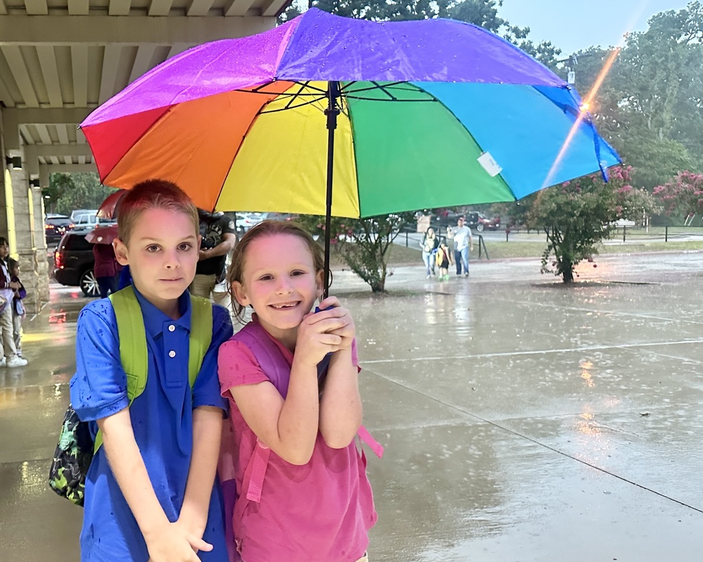 A young girl and boy, probably siblings, stand in front of a campus holding an umbrella while it's raining.