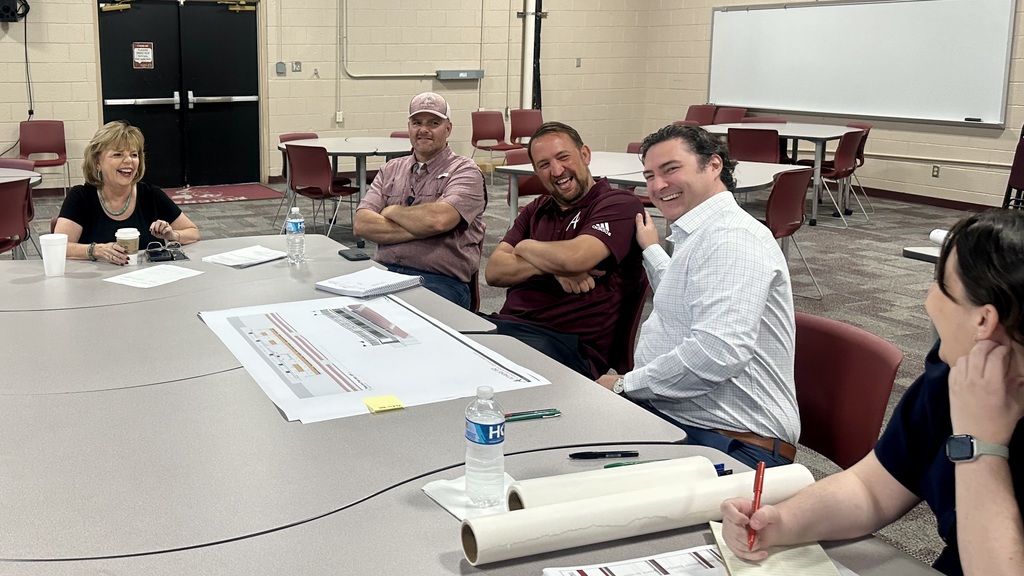Athens ISD staff and administrators share a lighthearted moment with representatives from Claycomb Associates Architects during a planning and design meeting in the Staff Development Room at the AISD Administration building. Five participants sit around a table with architectural drawings, notebooks, and drinks, smiling and laughing together. The background shows additional tables, chairs, and a whiteboard.