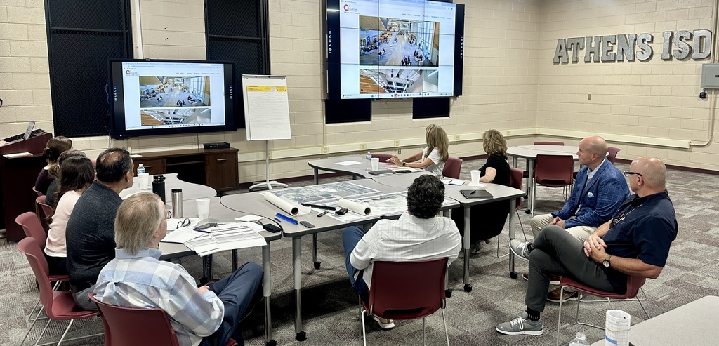 Athens ISD staff and administrators meet with representatives from Claycomb Associates Architects in the Staff Development Room at the AISD Administration building. Participants sit around tables with building plans, notebooks, and drinks, facing two large display screens showing architectural renderings of a modern school interior. A flip chart stands between the screens, and the room has “ATHENS ISD” lettering on the wall.