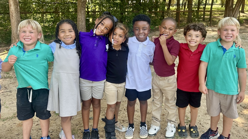 A group of kindergarten students at Bel Air Elementary stand together outdoors on the playground during recess, smiling and posing with arms around each other; they wear colorful school uniforms in shades of teal, gray, purple, black, white, burgundy, and red, with khaki or black shorts and skirts, and the background shows trees and a wooden fence.