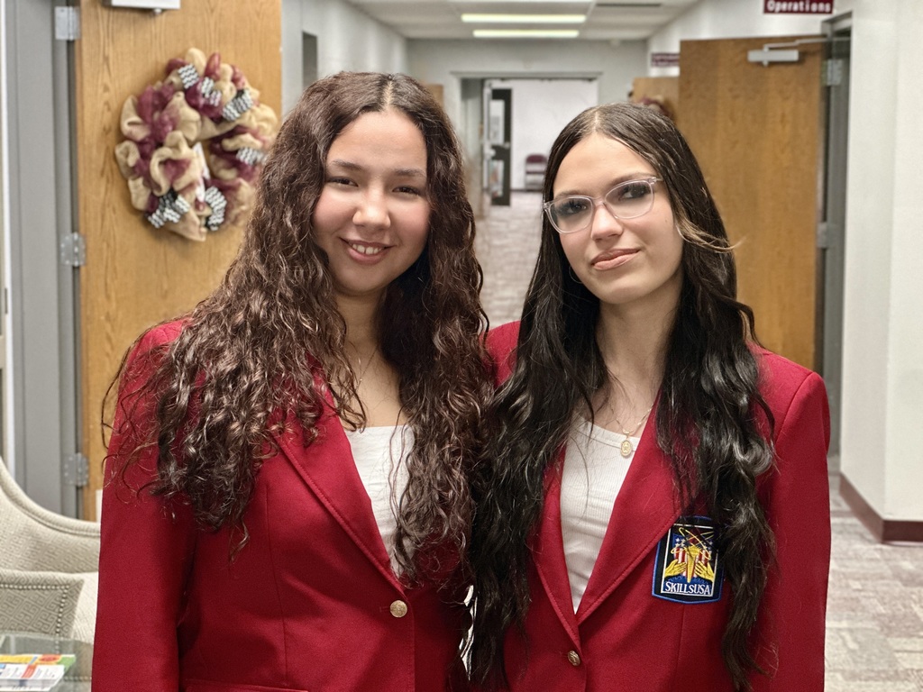 Two young women stand side by side indoors, both wearing red blazers over white tops. One has long curly brown hair and is smiling, while the other has long straight dark hair, wears glasses, and has a neutral expression. The woman on the right has a SkillsUSA patch on her blazer. The background shows open wooden doors and a hallway.