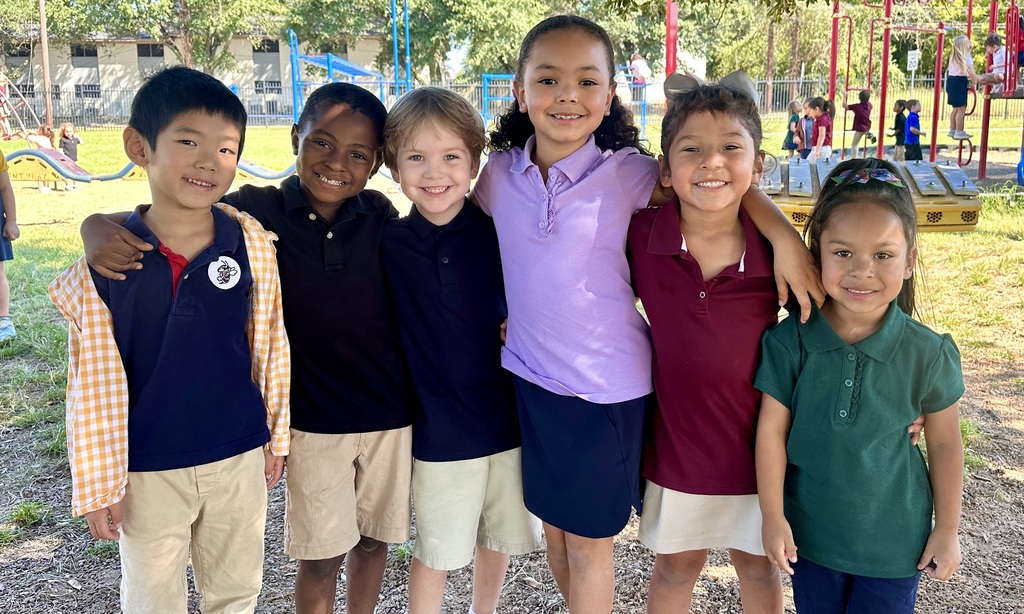 A group of six kindergarten children stands close together on the playground at Central Athens Elementary, smiling brightly with their arms around each other. They are dressed in colorful school uniforms, including navy, burgundy, green, and purple polo shirts paired with khaki or dark bottoms. The playground equipment and other children playing are visible in the background under the shade of trees.