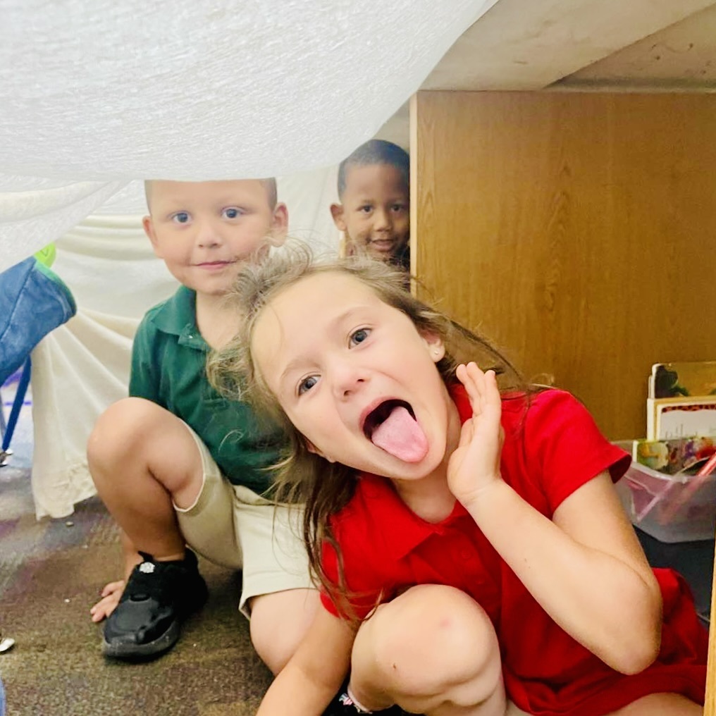 Three children are shown underneath what appears to be a sheet draped across a table or desk. They are smiling