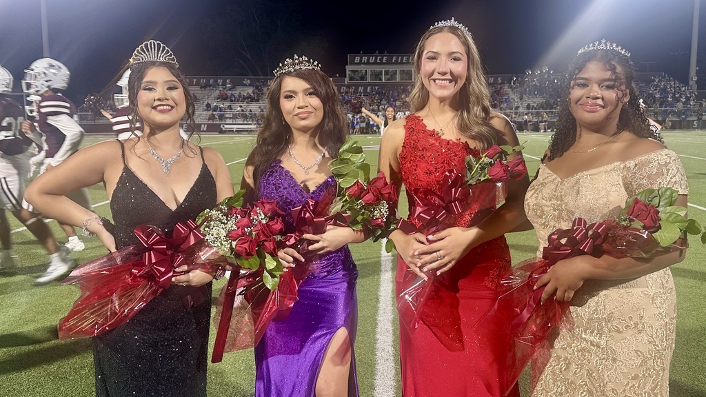 Four young women in formal gowns and tiaras stand side by side on a football field at night, each holding bouquets of red roses wrapped in red ribbon. They are smiling for the camera, with spectators and players visible in the background.