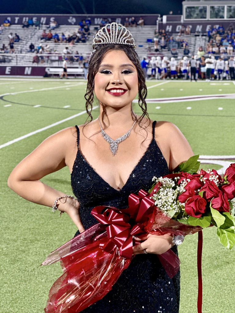 A young woman in a black gown and tiara stands on a football field, holding a bouquet of red roses wrapped in red ribbon. She is smiling brightly, wearing a sparkling necklace and bracelet.