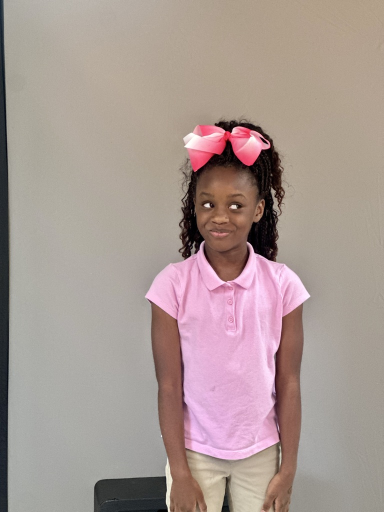 A young girl with a playful expression stands in front of a neutral gray backdrop. She wears a light pink polo shirt and khaki pants, with her hands relaxed at her sides. Her hair is styled in curls with a large pink and white bow on top. She glances to the side with a slight smirk, giving off a mix of curiosity and amusement.