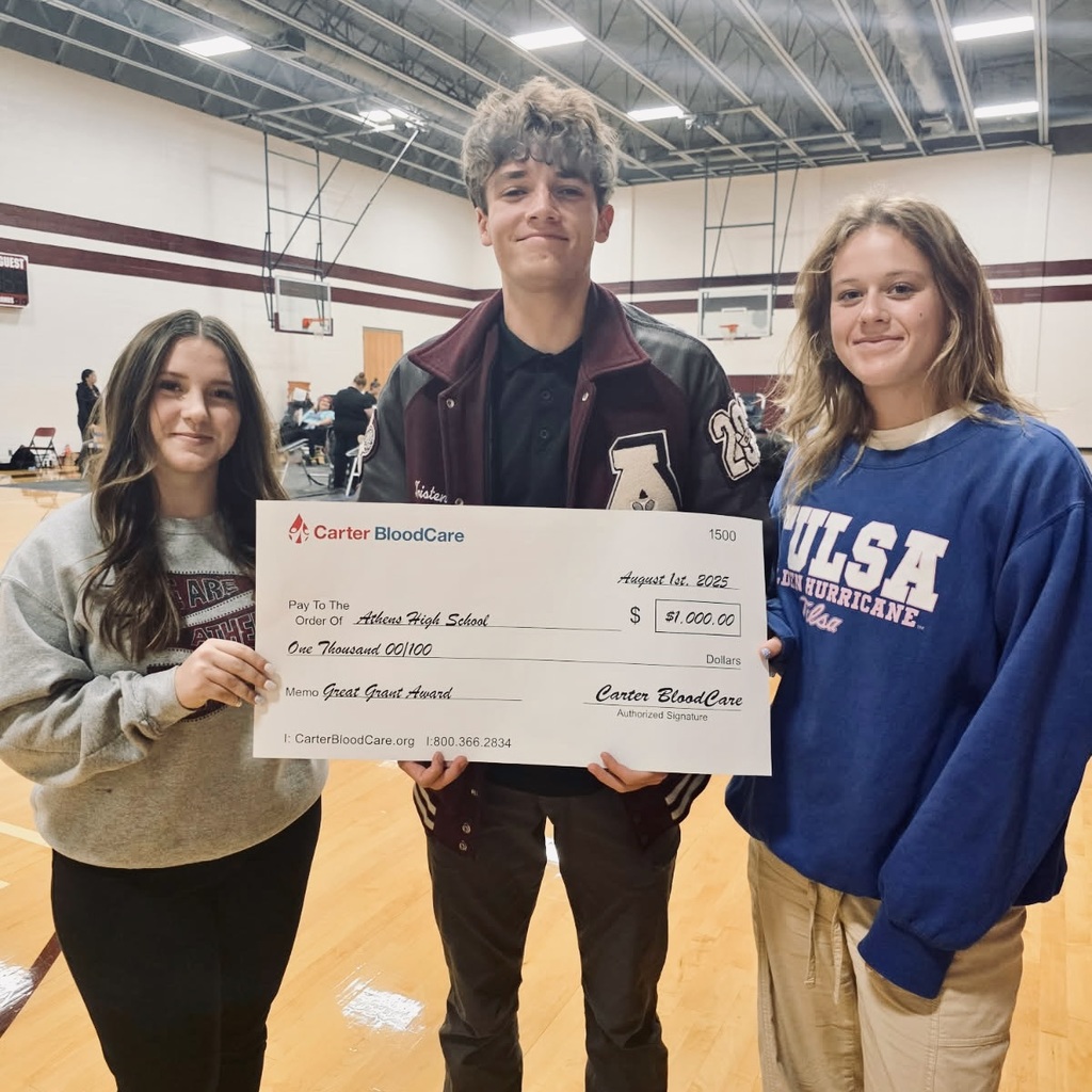 Three Athens High School students stand in a gym holding an oversized check from Carter BloodCare made out to Athens High School for $1,000. All three are smiling at the camera, and the check recognizes the school for reaching its blood donation goal. The students are identified as Sage Nelson, Tristen Husband, and Cobee Garner.