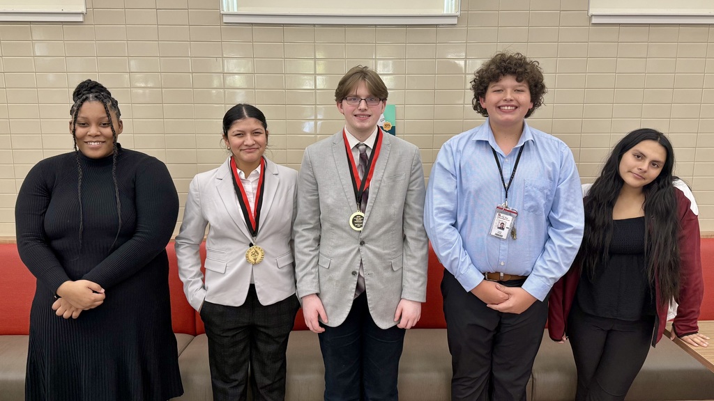 Five Athens High School debate students stand side by side and smile for a group photo indoors. Two students in the center wear medals around their necks. The group is standing in front of a tiled wall and sitting bench, dressed in a mix of professional and semi-casual attire.