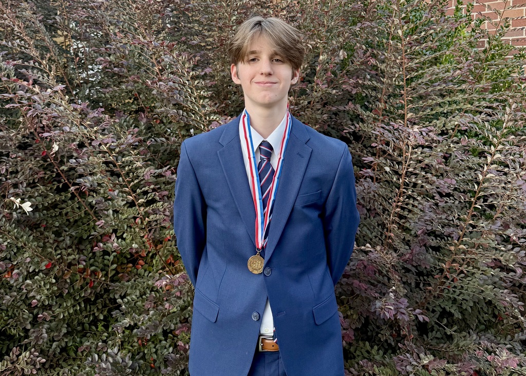 Liam Tinsley, an Athens High School junior, stands in front of leafy bushes wearing a blue suit, striped tie, and a single UIL medal around his neck, smiling confidently after earning a spot as an alternate to the state tournament in Congressional Debate.