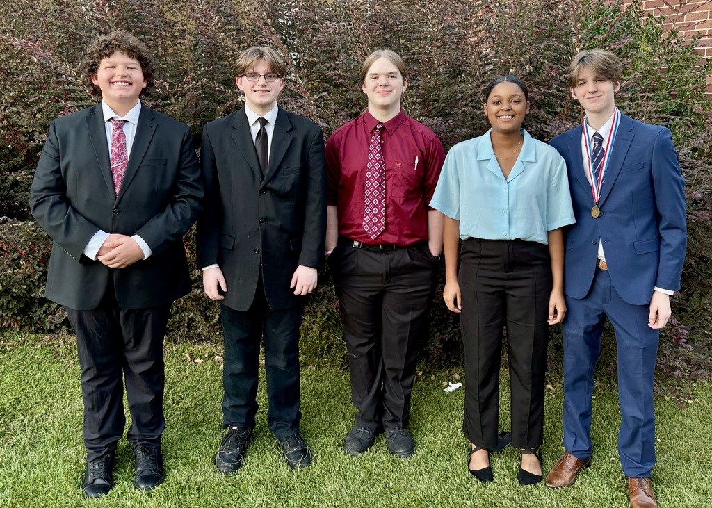 Five teenage students — four boys and one girl — are dressed formally or semi-formally, standing in front of foliage and smiling for the camera. One wears a medal.