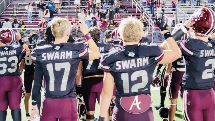 Photo of several football  players on the field taken from behind them as they lift their hands in celebration