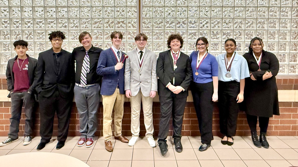 Nine Athens High School debate students stand in a row, smiling in front of a glass brick wall. Several students wear medals around their necks, and one holds a small gavel, celebrating their success at the Whitehouse Invitational speech and debate tournament.