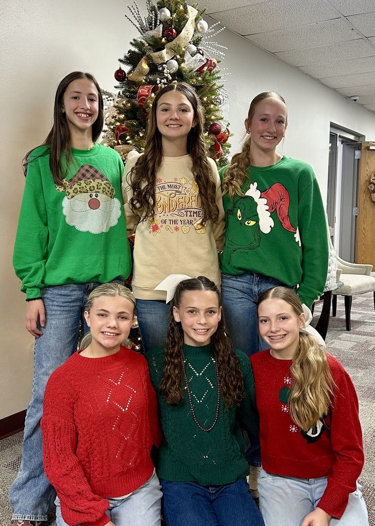 Six AMS Junior Honor Society members pose in front of the Christmas tree they decorated at the administration building. Three girls stand in the back row wearing festive holiday sweaters, and three girls kneel in front, also dressed in red and green sweaters. The decorated tree behind them features ribbon, ornaments, and lights.