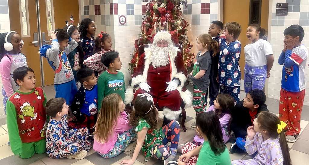 A group of excited second-grade students in colorful holiday pajamas gather around Santa Claus, who is seated in front of a decorated Christmas tree in a school hallway. The children are smiling, laughing, and looking toward Santa as he engages with them.