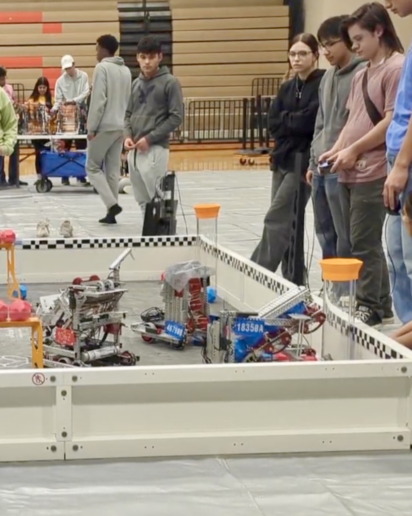Three Athens High School robotics team members stand at the edge of a competition field during a VEX match, focused on operating their robot. One student holds a controller while the others observe. The event takes place in a school gym with bleachers and tournament equipment in the background.