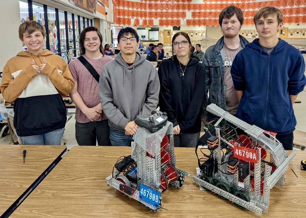 **Revised Alt Text:**  Six Athens High School robotics team members pose indoors behind two VEX V5 competition robots on a table. From left to right: Lyrick Adley, Wyatt McDonald, Josue Govea, Starlette Johnson, Brady Mayne, and Gaden Riquelme. The robot on the left has a blue license plate labeled "46790B" and the one on the right has a red plate labeled "46790A." Orange banners hang in the background of the school cafeteria setting.