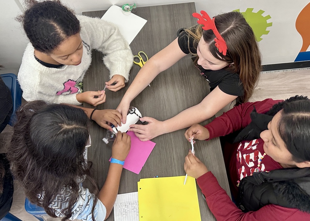 Overhead view of four elementary girls gathered around a table, working together on a STEM project. The students are using their hands to assemble materials around a small bottle, with scissors, paper, and notebooks spread across the table, showing teamwork and hands-on problem-solving.